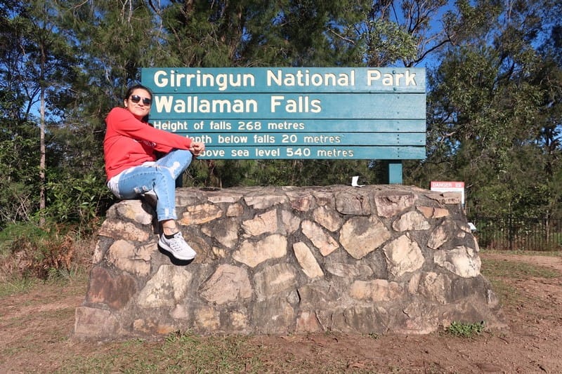 Vivir en Brisbane chica morena con chaqueta roja y tejanos sentada en un muro con un cartel detrás
